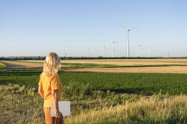 Woman holding laptop looking at wind turbine in field - SVKF00413