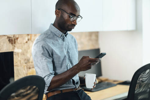 Geschäftsmann mit Kaffeebecher und Mobiltelefon am Arbeitsplatz - JSRF02166