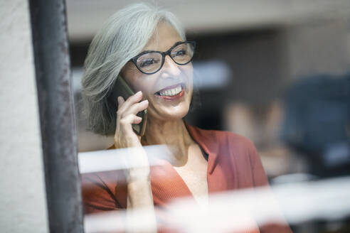 Lächelnde ältere Geschäftsfrau am Telefon durch ein Glasfenster gesehen - JSRF02168