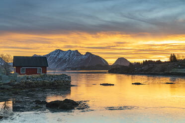 Schneebedeckte Berge und rote Fischerhütten unter dem brennenden Himmel im Morgengrauen, Ballstad, Vestvagoy, Lofoten, Norwegen, Skandinavien, Europa - RHPLF22571