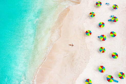 Luftaufnahme einer Frau im Bikini beim Sonnenbaden an einem tropischen Strand neben bunten Sonnenschirmen, Antigua, Westindien, Karibik, Mittelamerika - RHPLF22581