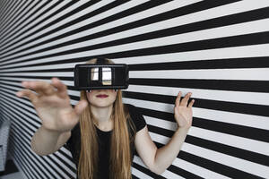 Teenager-Mädchen gestikuliert mit Virtual-Reality-Headset vor gestreifter Wand - LLUF00814