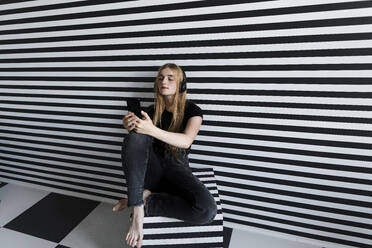 Teenage girl with headphones using smart phone sitting by striped wall - LLUF00818