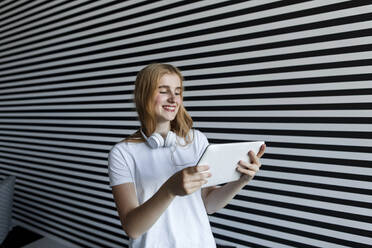 Smiling beautiful girl with headphones watching tablet PC by striped wall - LLUF00823