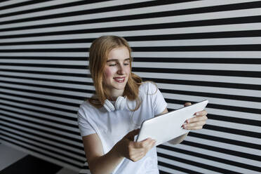 Beautiful teenage girl using tablet PC by striped wall - LLUF00825