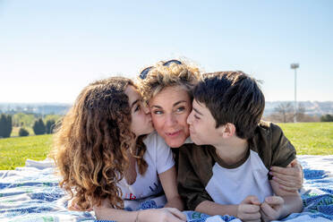 Geschwister, die ihre Mutter küssen, während sie auf einer Picknickdecke gegen den Himmel an einem sonnigen Tag liegen - ADSF36101