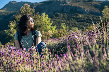 Ernste asiatische Wanderin, die auf einer Wiese mit blühenden lila Wildblumen sitzt, während eines Trekkings in der Natur an einem Sommertag in Valverde de los Arroyos - ADSF36122