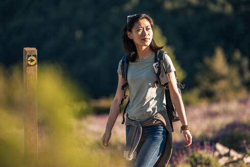 Seitenansicht einer asiatischen Wanderin in Freizeitkleidung mit Rucksack, die beim Trekking an einem sonnigen Sommertag in Valverde de los Arroyos in der Natur mit grünen Pflanzen spazieren geht - ADSF36123