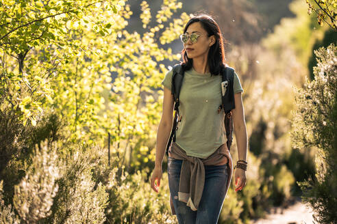 Asiatische Wanderin in Freizeitkleidung und Sonnenbrille mit Rucksack spaziert in der Natur mit grünen Pflanzen während des Trekkings an einem sonnigen Sommertag in Valverde de los Arroyos - ADSF36125