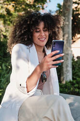 Positive hispanische Unternehmerin im weißen Anzug mit lockigem Haar, die lächelt und ein Selfie mit ihrem Smartphone macht, während sie an einem sonnigen Tag im Park auf einer Bank sitzt - ADSF36134