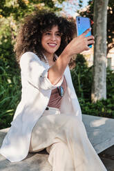 Positive hispanische Unternehmerin im weißen Anzug mit lockigem Haar, die lächelt und ein Selfie mit ihrem Smartphone macht, während sie an einem sonnigen Tag im Park auf einer Bank sitzt - ADSF36135