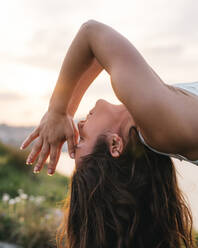 Seitenansicht einer ruhigen jungen Frau mit langen braunen Haaren, die eine Rückbeuge-Asana mit geschlossenen Augen und betenden Händen in der Nähe des Gesichts macht, während sie Yoga am Meer bei Sonnenuntergang praktiziert - ADSF36143