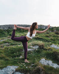 Side view of concentrated young female with long brown hair in sportswear standing on grassy hill in Dandayamana Dhanurasana pose while practicing yoga in highlands - ADSF36144