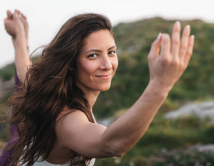 Positive junge Frau mit langen braunen Haaren, die lächelt und in die Kamera schaut, während sie während einer Yoga-Sitzung in einem Gebirgstal eine stehende Verbeugung (Asana) ausführt - ADSF36145