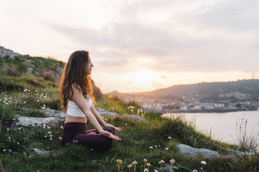 Seitenansicht einer fitten Sportlerin in Sportkleidung, die auf einem Grashügel in Padmasana-Pose sitzt und bei Sonnenuntergang Yoga praktiziert - ADSF36146