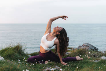 Full body of female athlete leading healthy lifestyle and practicing yoga while sitting on grassy seashore in Ardha Kapotasana pose with hand in gyan mudra and looking up - ADSF36147