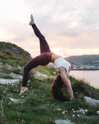 Junge Sportlerin in Sportkleidung in Urdhva Dhanurasana-Pose auf grasbewachsenem Meeresufer in voller Länge - ADSF36148