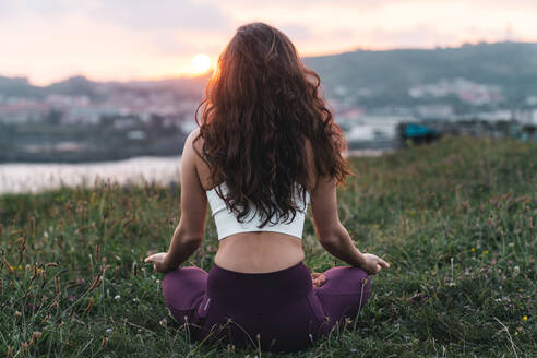 Rückenansicht einer fitten Sportlerin in Sportkleidung, die auf einem Grashügel in Padmasana-Pose sitzt und bei Sonnenuntergang Yoga praktiziert - ADSF36149