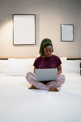 Full body of young ethnic female sitting on bed and browsing netbook while working remotely in modern bedroom at home - ADSF36167