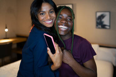 Fröhliche multirassische junge Frauen mit Smartphone lächelnd und umarmen einander, während stehend in der Nähe von Bett in schummrigen Schlafzimmer am Abend zu Hause - ADSF36172