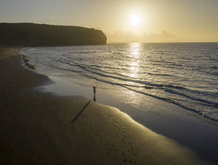 Luftaufnahme einer Frau, die bei Sonnenuntergang am Strand entlang läuft, Praia Areal de Santa Barbara, Ribeira Seca, Azoren, Portugal. - AAEF15161