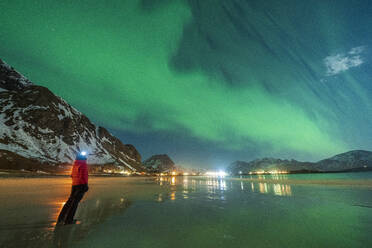 Hiker with head torch watching the Aurora Borealis (Northern Lights) standing on Ramberg beach, Nordland county, Lofoten Islands, Norway, Scandinavia, Europe - RHPLF22681