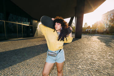 Woman with skateboard standing under bridge at sunset - MEUF07531
