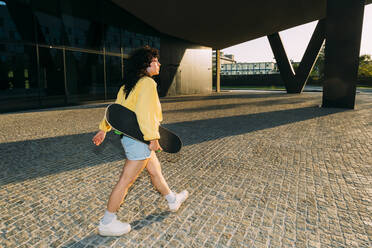 Woman walking with skateboard under bridge at sunset - MEUF07532
