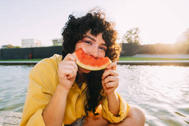 Young woman eating watermelon by pond at sunset - MEUF07542