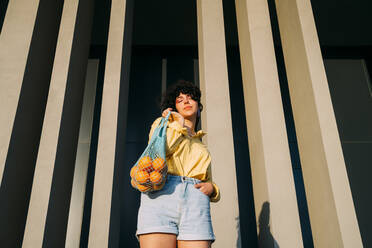 Woman with eyes closed carrying oranges in mesh bag in front of column - MEUF07545