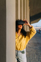 Woman wearing virtual reality simulator by column on footpath - MEUF07567