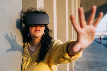 Young woman with VR glasses doing stop gesture by column - MEUF07569