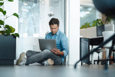 Businessman using laptop sitting on floor in office - KNSF09565