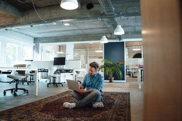 Geschäftsmann mit Laptop sitzt auf Teppich im Büro - KNSF09569