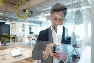 Älterer Geschäftsmann, der mit seinem Mobiltelefon im Internet surft, gesehen durch Glas im Büro - KNSF09580