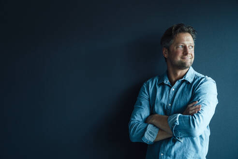 Smiling businessman standing with arms crossed against blue background - KNSF09597