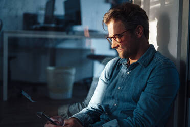 Smiling businessman wearing eyeglasses using smart phone sitting in office - KNSF09601