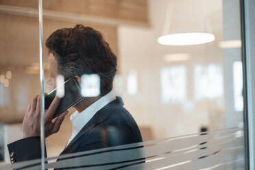 Businessman talking on smart phone seen through glass in office - KNSF09611