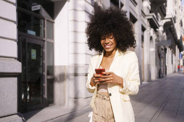 Lächelnde Frau mit lockigem Haar, die ein Smartphone benutzt und auf dem Bürgersteig geht - MMPF00193