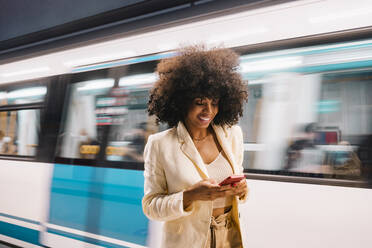 Smiling woman using smart phone in front of moving subway train - MMPF00218