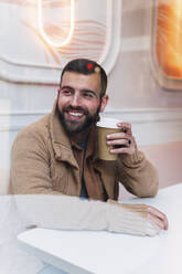 Happy young man with disposable coffee cup sitting at table in cafe - PNAF04308