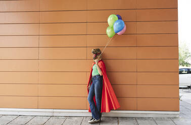 Frau mit Krokodilmaske lehnt mit bunten Luftballons an der Wand - JCCMF06886