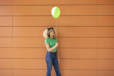 Junge Frau mit grünem Luftballon vor einer Wand stehend - JCCMF06894