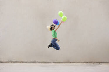 Glückliche junge Frau mit bunten Luftballons, die vor einer Wand springen - JCCMF06901