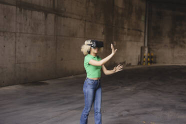 Young woman wearing virtual reality simulator gesturing in parking garage - JCCMF06904