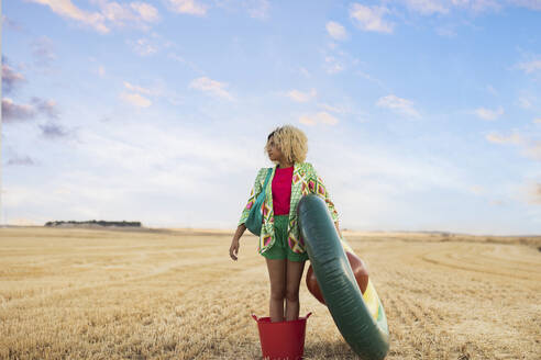 Young woman standing with avocado shaped inflatable ring at field - JCCMF06908