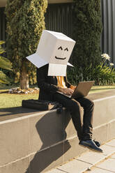 Businessman wearing box with smiley face using laptop sitting on wall - MGRF00700