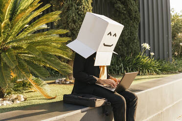 Freelancer wearing box with smiley face using laptop sitting on wall - MGRF00701