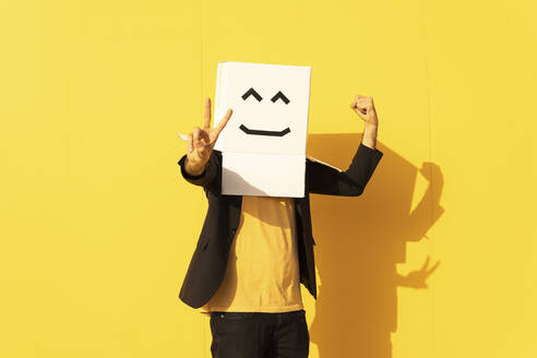 Mann trägt Schachtel mit Smiley-Gesicht und gestikuliert Friedenszeichen vor gelber Wand - MGRF00716