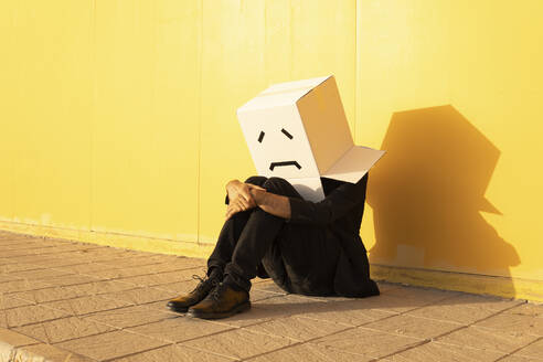 Man wearing box with sad face sitting on footpath - MGRF00726